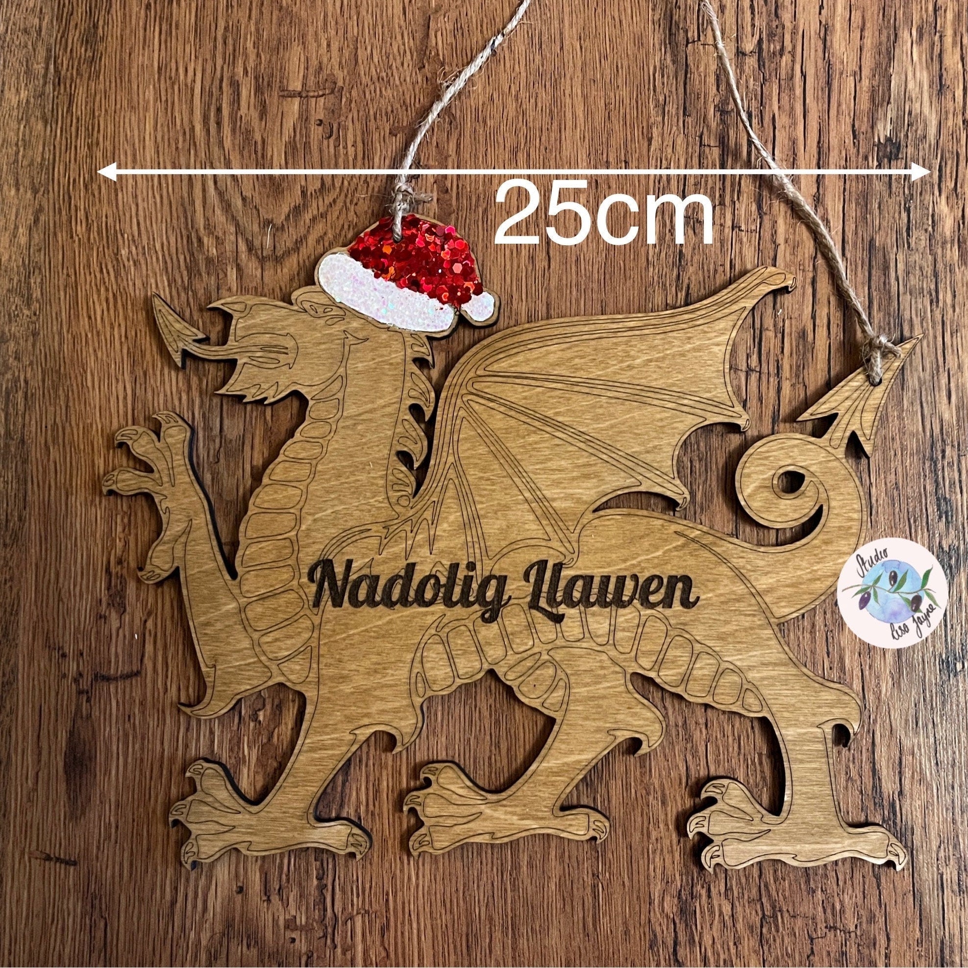 A wooden hanging sign featuring a red Welsh dragon with a Santa hat, the text 'Nadolig Llawen' carved in the center, and decorated with red and silver glitter. The sign is 25cm wide.
