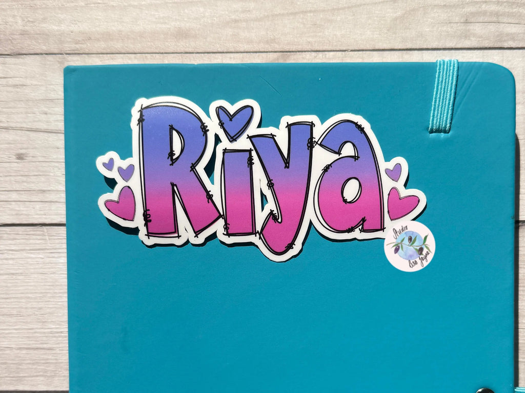 Blue diary notebook with 'Riya' Ombré Heart Name Personalised Sticker in colorful letters on a wooden surface