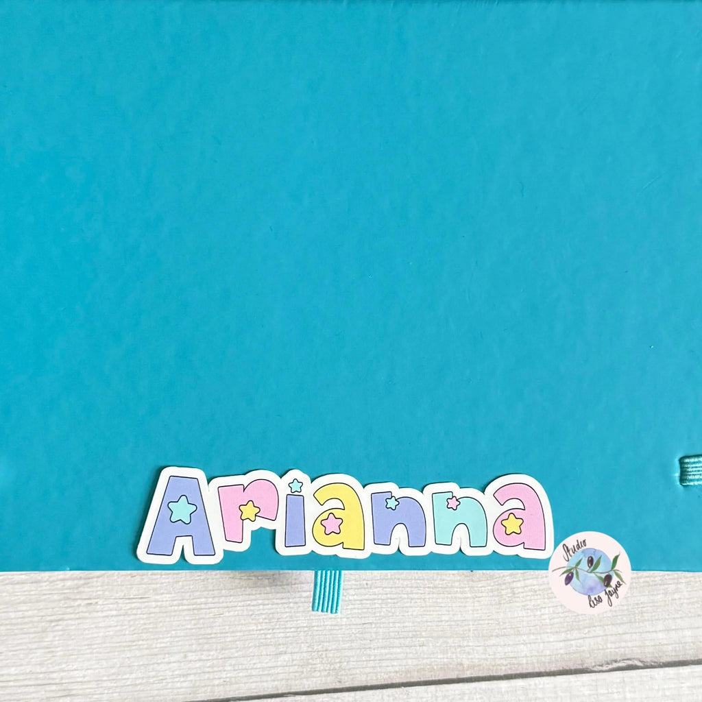 Turquoise notebook diary with colorful name 'Arianna' and a sticker on a wooden surface