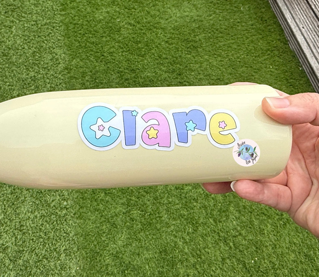 Hand holding a water drink bottle with 'Clare' in colorful pastel star letters on a grassy background