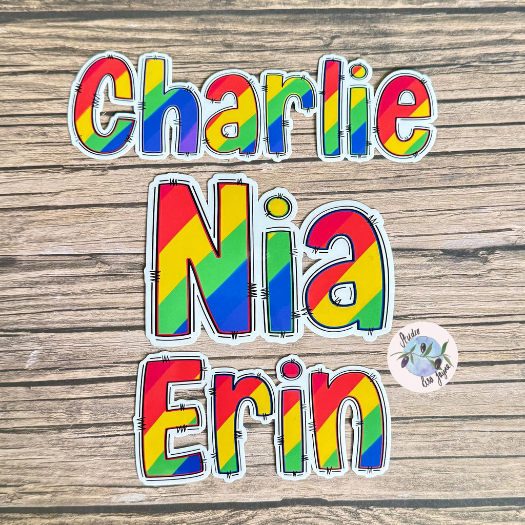 Colourful rainbow name stickers on a wooden surface with a small sticker in the corner.