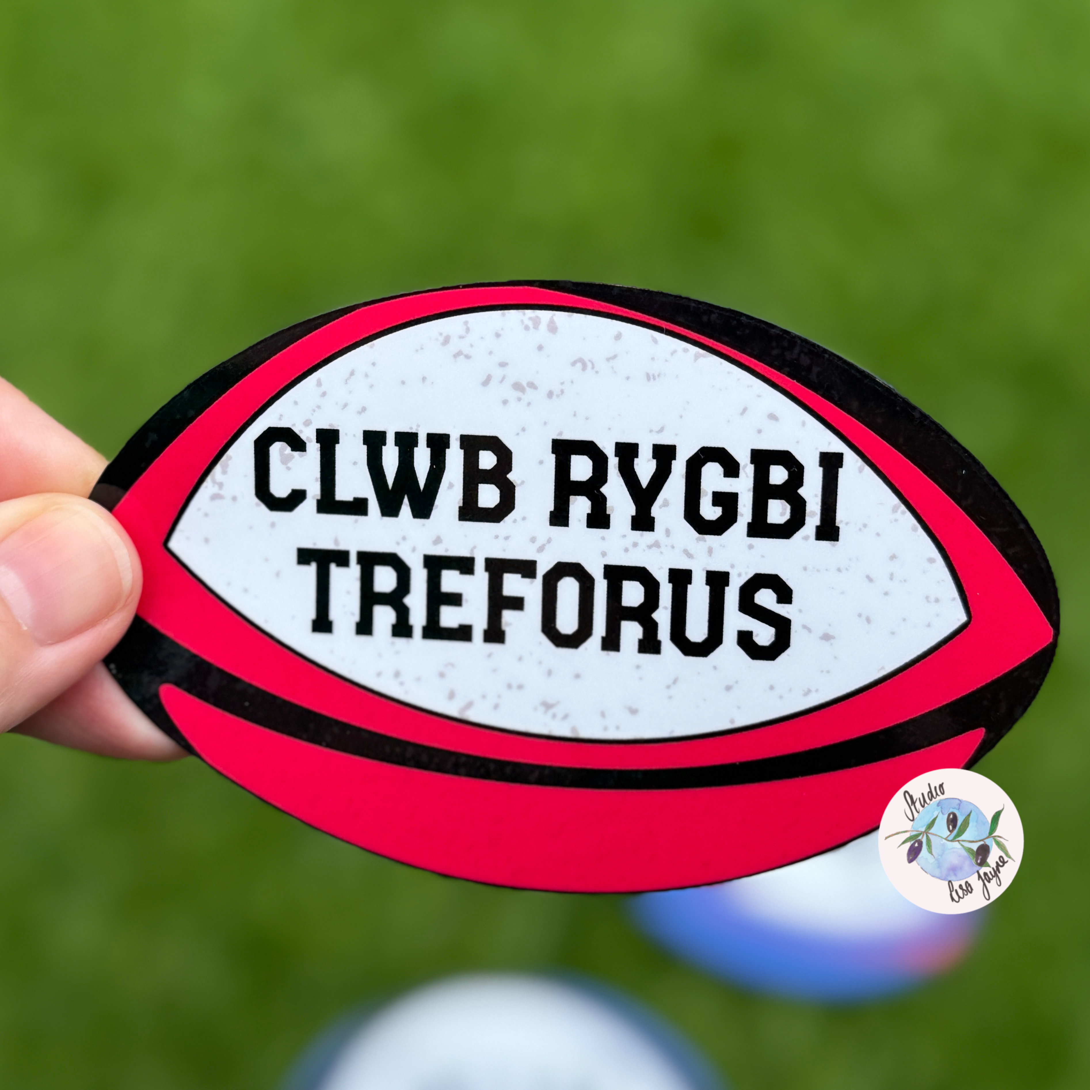 Red and white rugby ball-shaped sticker with 'CLWB RYGBI TREFORUS' text on a green background.  Ideal for your local rugby team