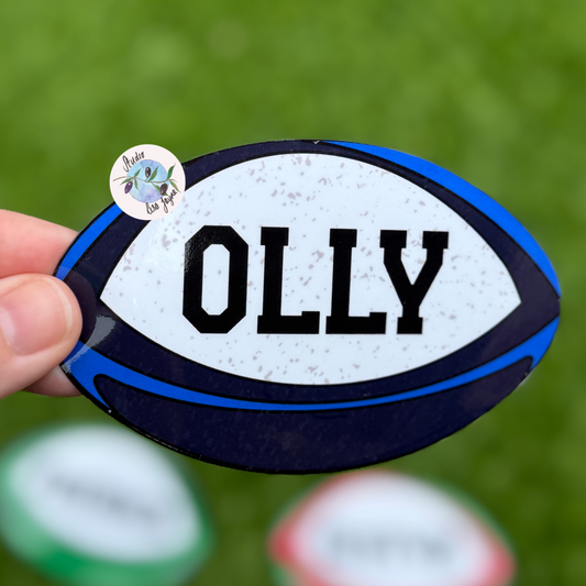 Scottish rugby ball sticker with 'OLLY' name printed on it, held by a hand against a blurred green background.