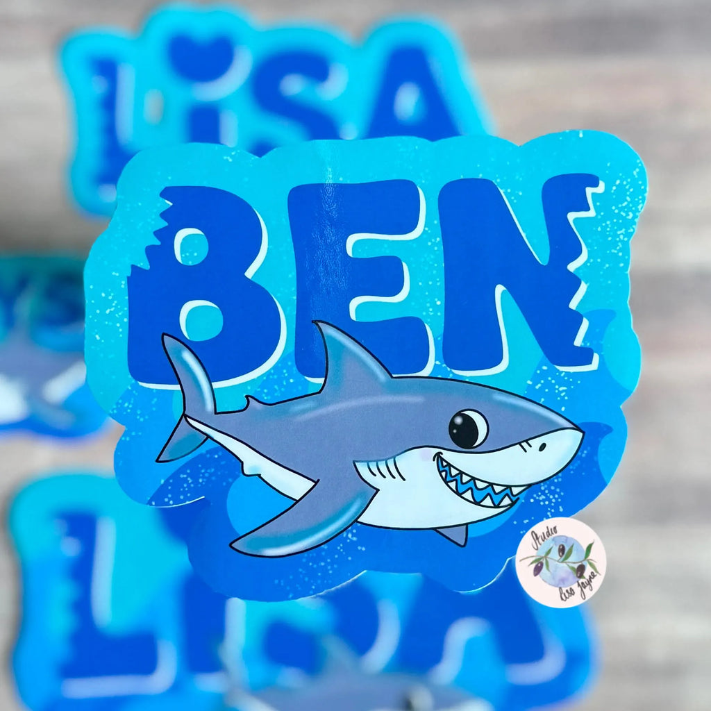 Blue name stickers with a shark design and personalized names on a wooden surface.