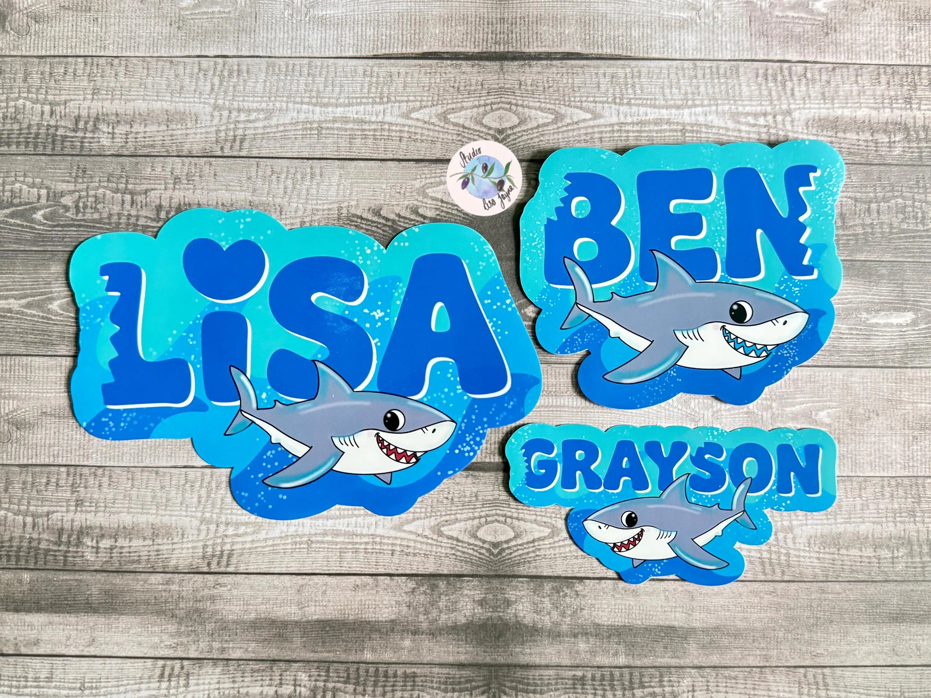 Shark-themed name vinyl decals for Lisa, Ben, and Grayson on a wooden surface.