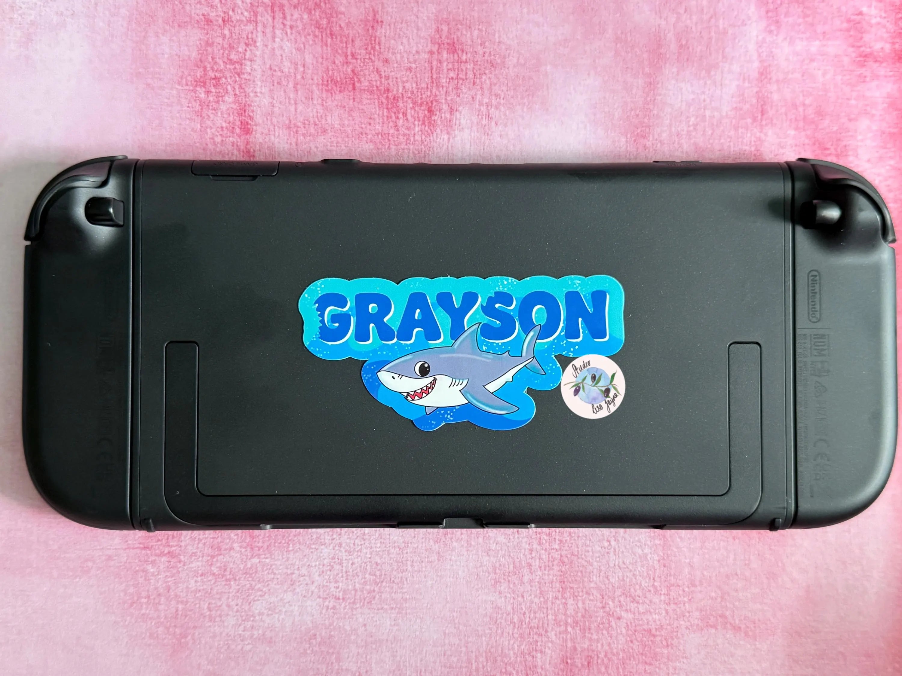 Black gaming console with a shark-themed 'Grayson' sticker on a pink textured background.  Nintendo Switch 2