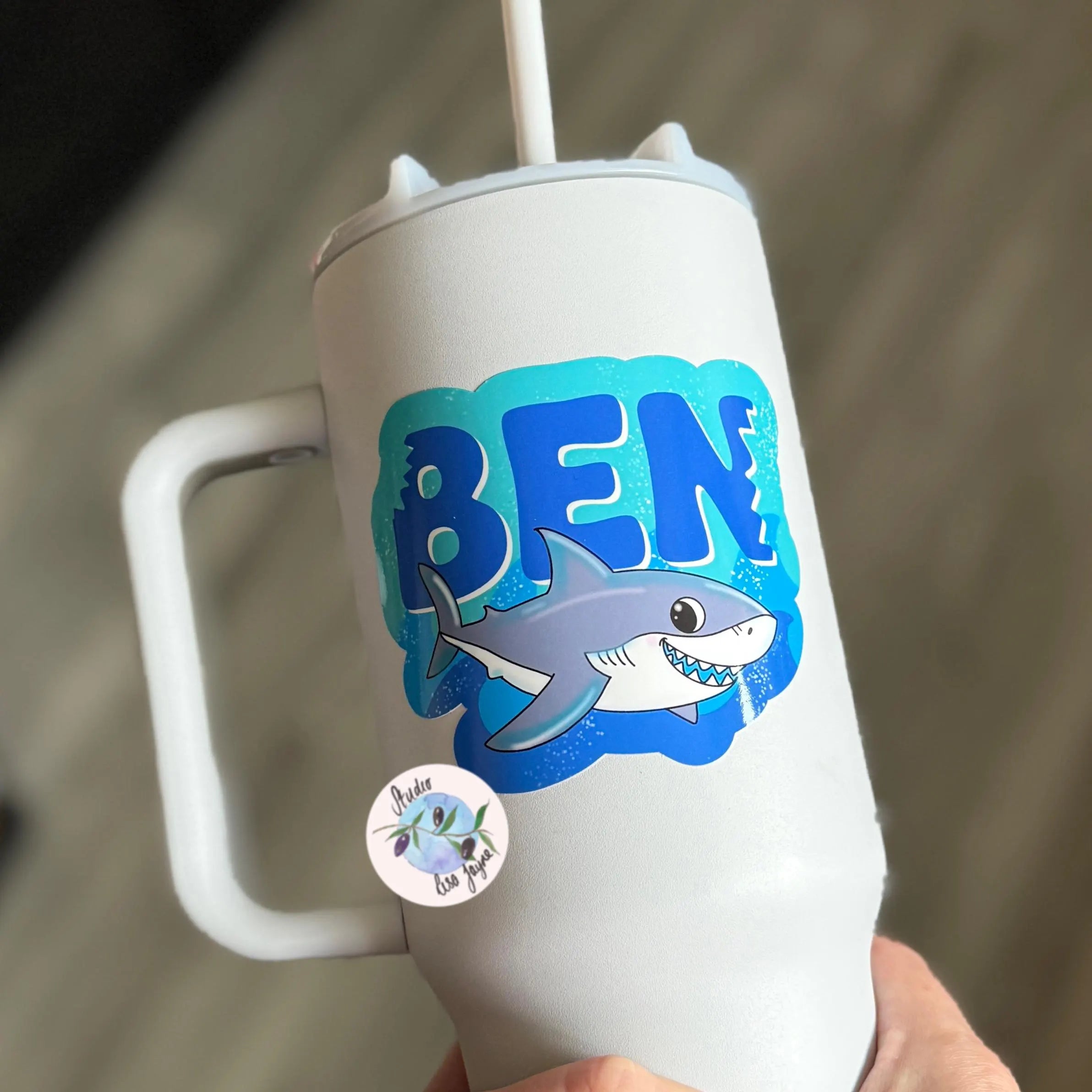 White tumbler with shark design vinyl label and 'BEN' text on a blurred background