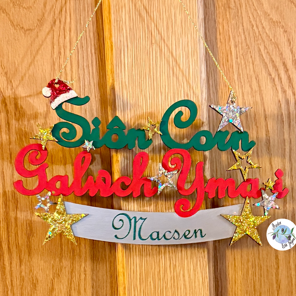 A personalised Christmas hanging sign with wooden letters spelling 'Sion Con Galwch Yma i' and a banner reading 'Macsen', decorated with gold glitter stars, a red Santa hat on a child’s Welsh Christmas door, welcoming Santa