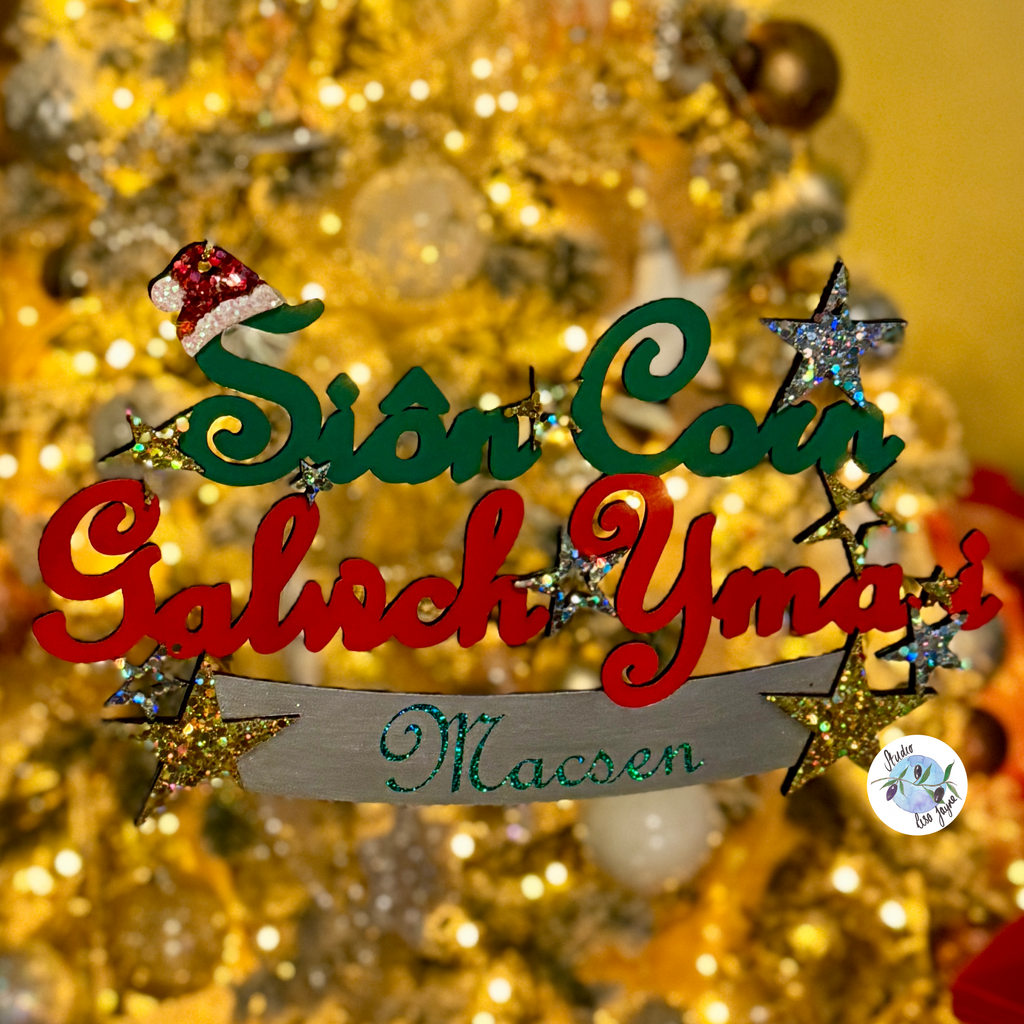 A decorative Christmas sign with the words 'Sion Coin Galwch Yma i Macsen' in red, green, and silver lettering, adorned with glittery stars and a small Santa hat, set against a blurred golden Christmas tree background.