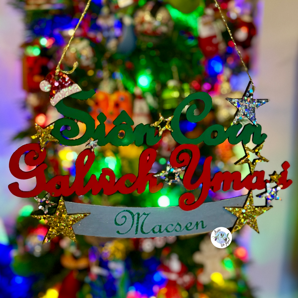 A personalised Christmas hanging sign with the text 'Siôn Corn Galwch Yma I ' in green and red letters, a silver banner reading 'Macsen' in green script, and gold glitter stars, hanging in front of a blurred, brightly lit Christmas tree.