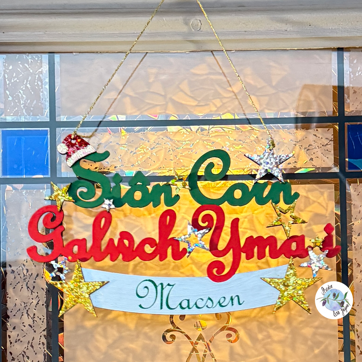 A festive hanging sign with the words 'Sion Corn Galwch Yma i' in red and green lettering, decorated with glittery gold stars, a Santa hat ornament, and a silver banner reading 'Macsen'. The sign is displayed on a window with a golden light background.

Perfect Christmas Welsh gift