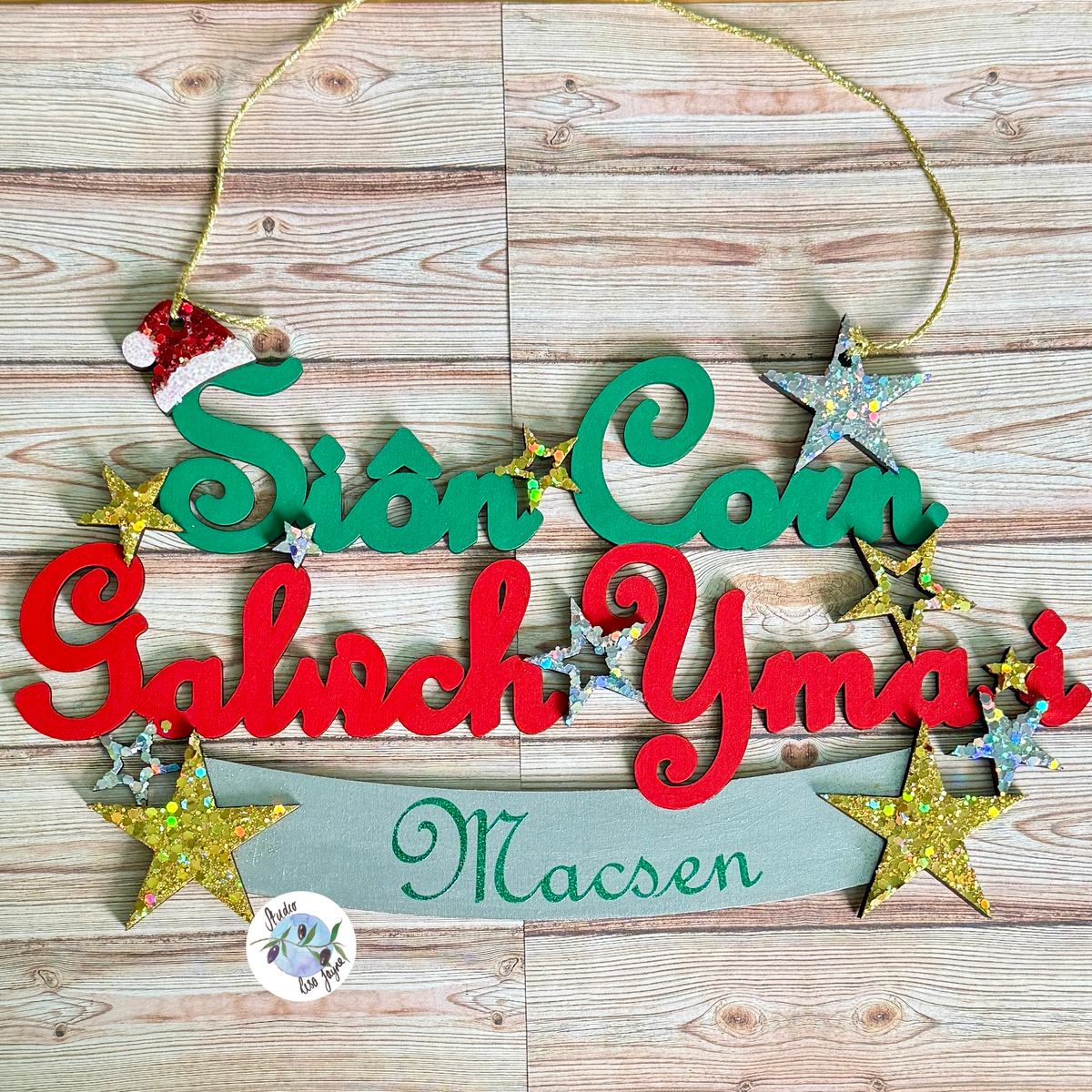 A personalised Christmas wooden sign with red and green lettering reading 'Sion Coin Galwch Yma i' and a banner below reading 'Macsen', decorated with gold glitter stars, a small Santa hat ornament, and a gold hanging string, shown on a light wood plank background.

Cymru Welsh Christmas Nadolig