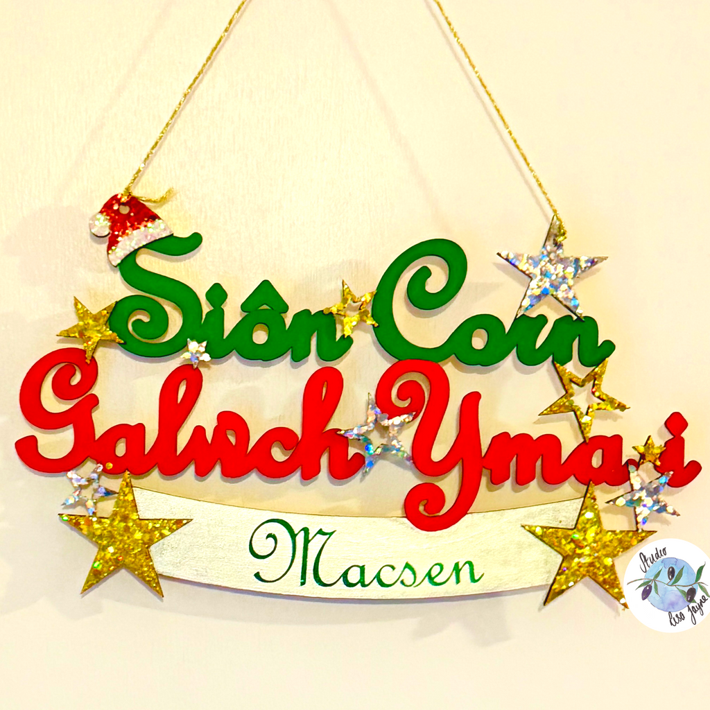 A personalised Christmas hanging sign with the text 'Sion Corn Galwch Yma i' in green and red letters, decorated with gold glitter stars, a small Santa hat ornament, and a cream banner reading 'Macsen' in green script. The sign hangs from a gold string against a light beige wall.