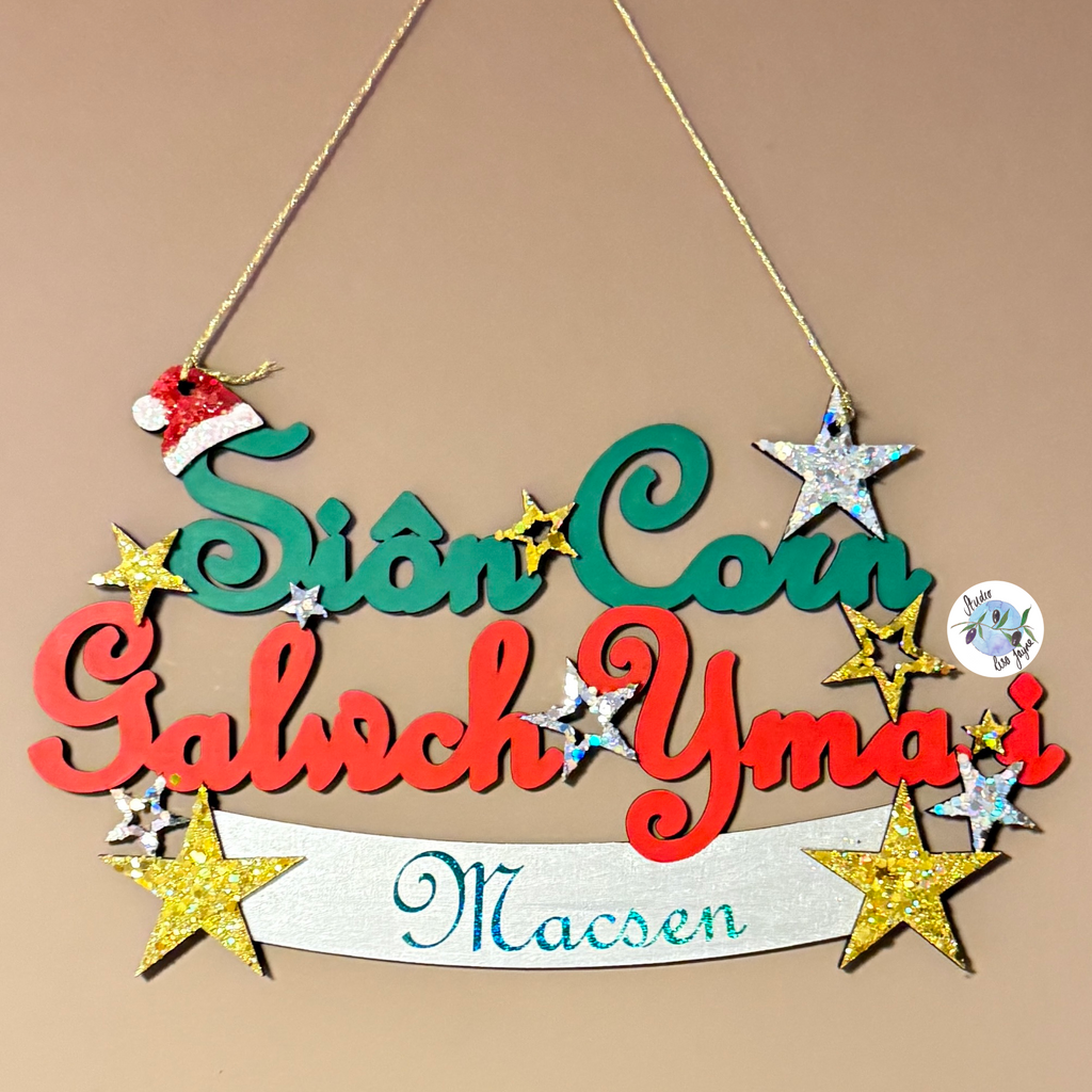 A personalised wooden Christmas hanging sign with the message 'Sion Corn, Galwch Yma i', and 'Macsen' in colourful script and block letters, decorated with glittery stars, a small Santa hat, and a white banner at the bottom, shown against a plain beige background.