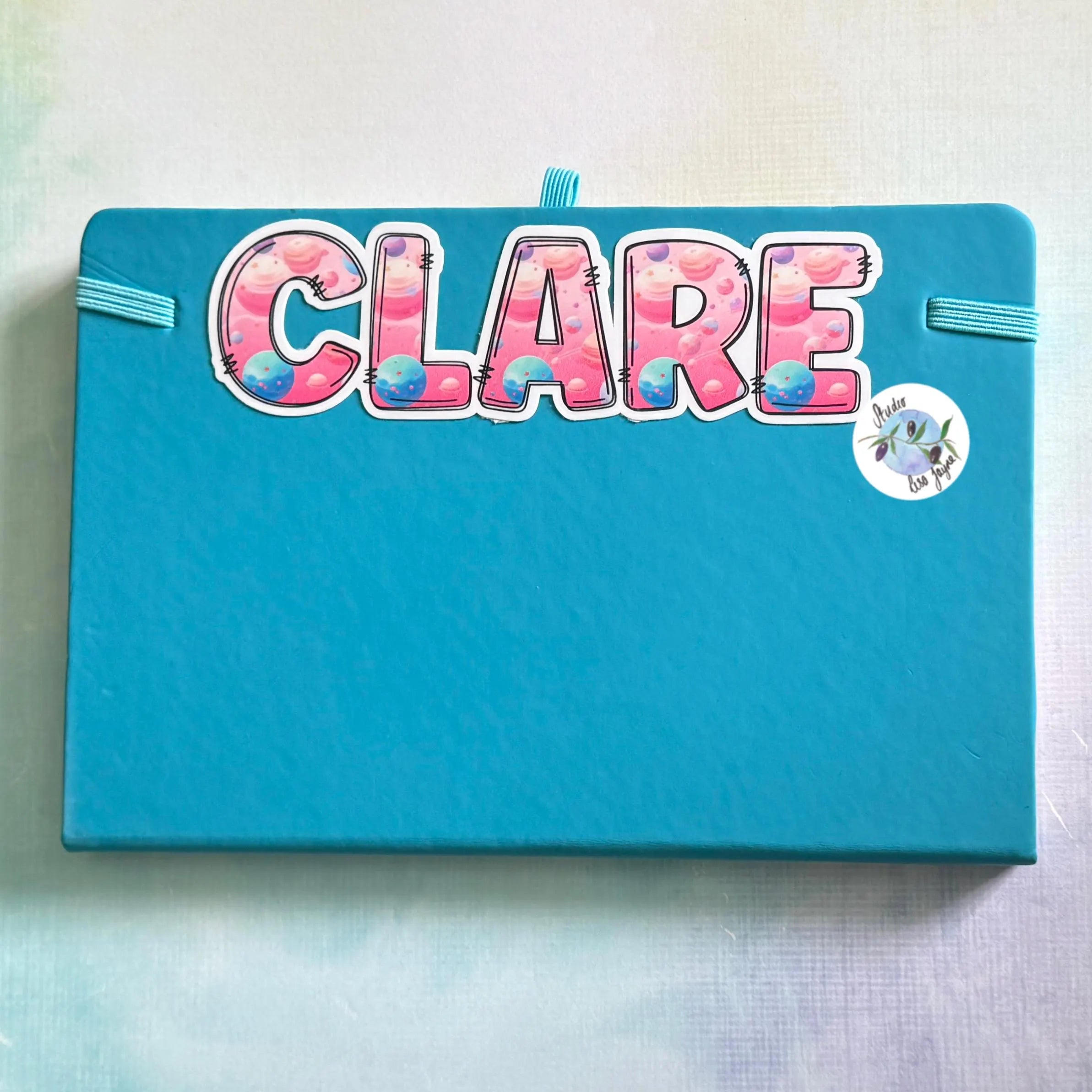 Blue notebook with 'CLARE' in colorful space themed letters on a vinyl decal sticker label