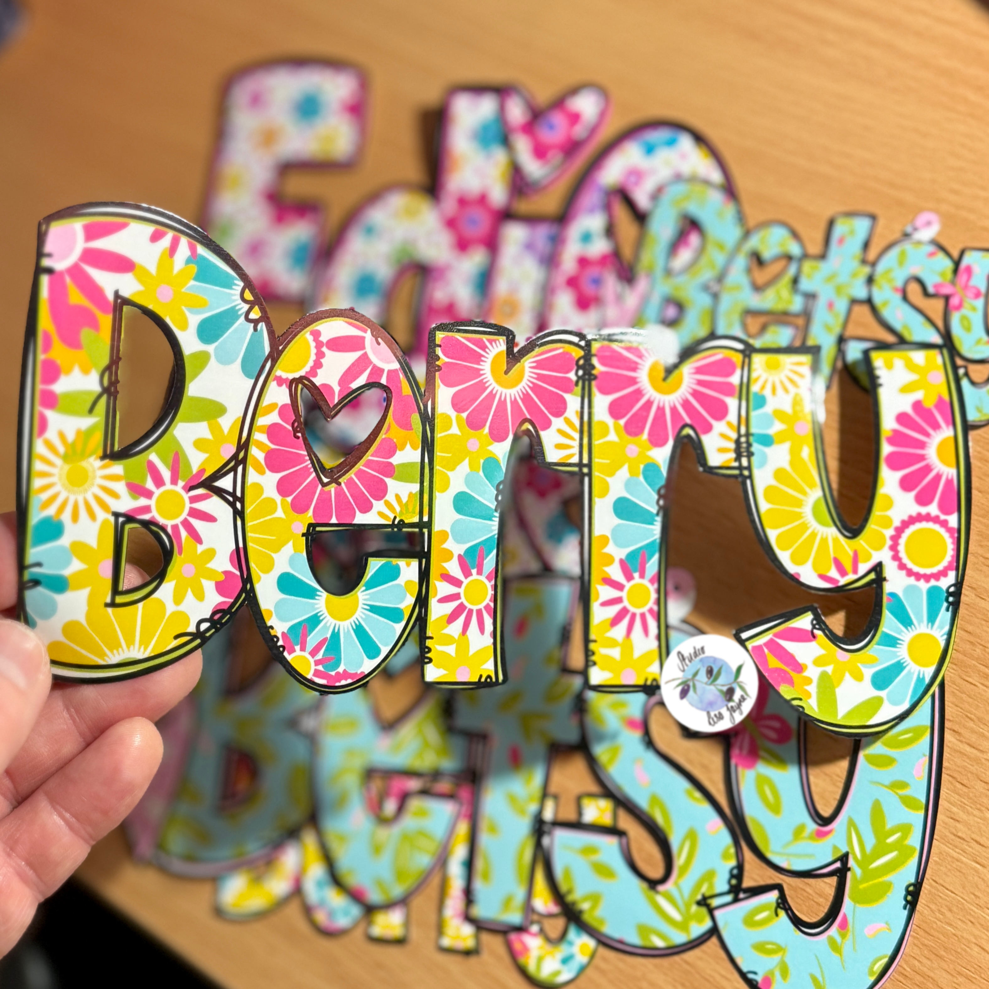 A hand holds a colourful name sticker spelling 'Berry' with a floral pattern, in front of matching cutouts on a wooden surface.