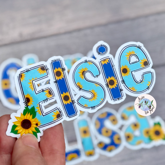 Sunflower Sticker with 'Elsie' text and held by a hand on a wooden surface