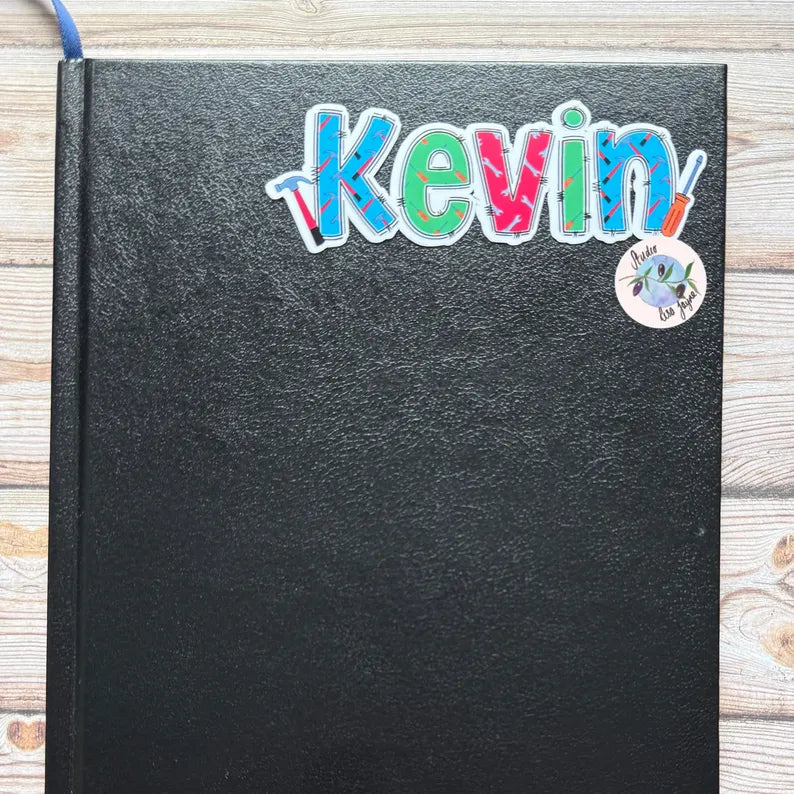 Black notebook diary with colorful tool themed name 'Kevin' and a sticker on a wooden surface