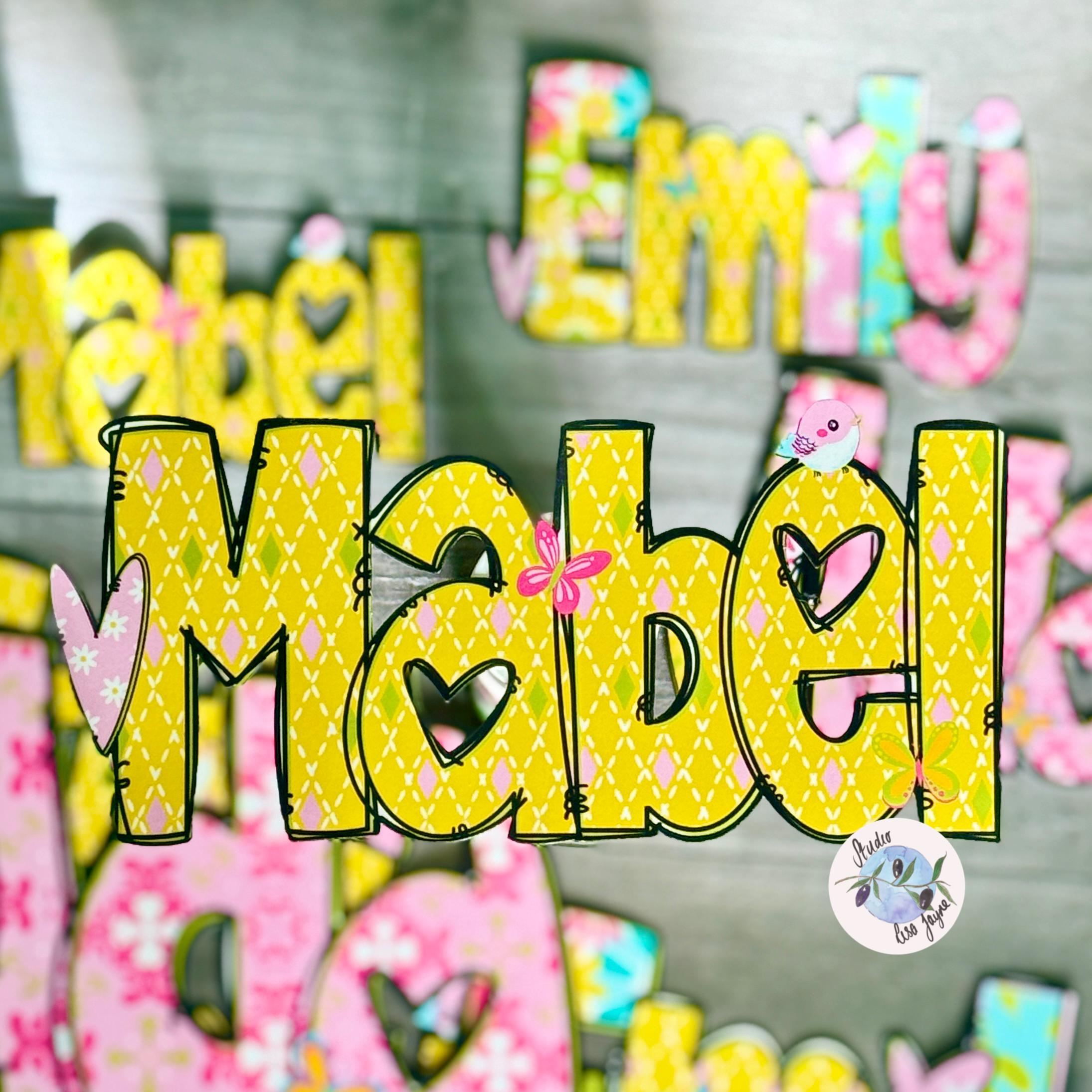 A close-up of decorative personalized lettering spelling names, with the largest and sharpest letters reading 'Mabel' in bright yellow with a small pink bow and heart details, alongside other colorful letters in pink, blue, and yellow on a gray background.