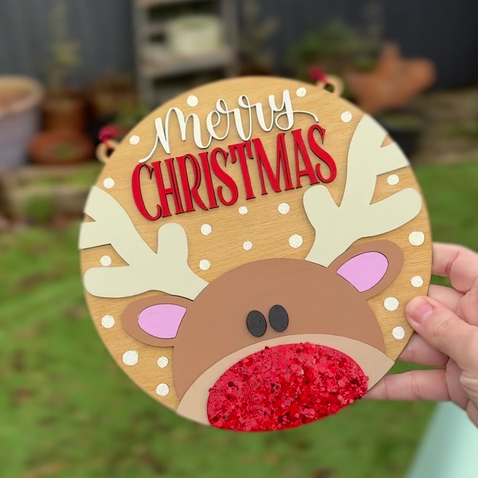 Wooden hand painted reindeer ornament with 'Merry Christmas' text on a green leafy background
